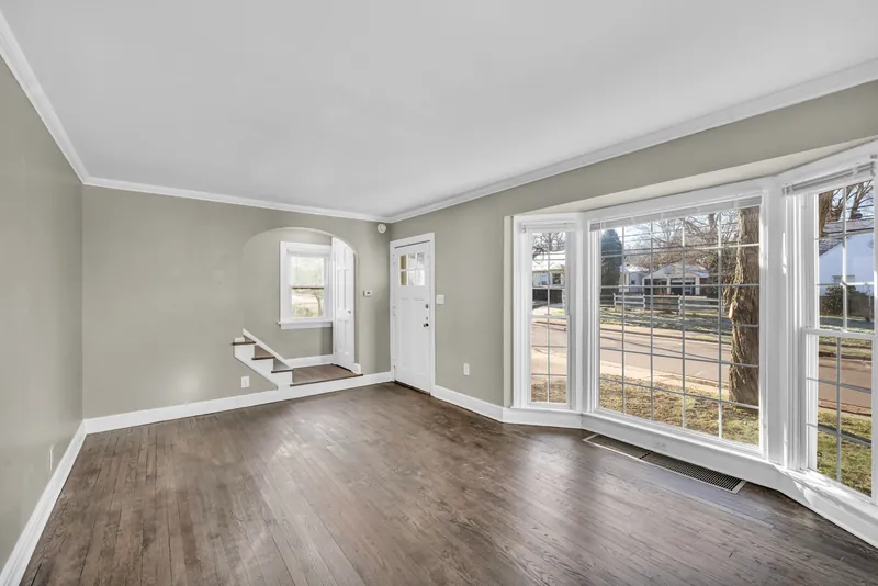 Before: empty living room with gray-beige walls, dark hardwood floors, white crown molding, a front door, and a large white bay window